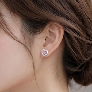 Könnte beinhalten: Ein herzförmiger Ohrstecker mit einem zentralen rosa Edelstein, der von einem Kranz aus kleinen, klaren, funkelnden Steinen in einer Metallfassung umgeben ist. Der Ohrstecker befindet sich am Ohrläppchen und zeigt ein klassisches Design.