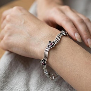 May include: A silver-toned bracelet featuring a multi-strand wavy design with oxidized detailing. The bracelet is accented with small, five-petaled flower motifs, each centered with a small, round, deep red gemstone set in the middle of the metal petals.