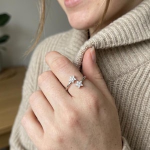Può includere: Un anello aperto color argento con due motivi a forma di stella alle estremità. Ogni stella è incastonata con piccole pietre chiare e scintillanti. La fascia è sottile e lucida, progettata per avvolgersi leggermente attorno al dito.