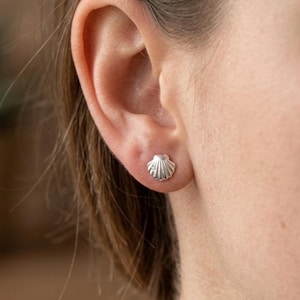 Peut inclure: Une petite boucle d'oreille à clou en argent en forme de coquillage avec une surface texturée en éventail et une minuscule pierre claire incrustée à la base, fixée au lobe. La boucle présente une finition métallique et un design strié.