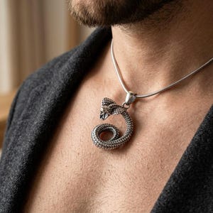 Op de afbeelding: Een zilverkleurige metalen hanger in de vorm van een opgerolde cobra met gedetailleerde schubben en een open mond, hangend aan een dunne, gladde zilverkleurige slangenketting die op het sleutelbeen rust.
