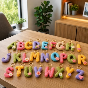 Könnte beinhalten: Eine Sammlung von sechsundzwanzig gehäkelten Buchstaben-Schlüsselanhängern, jeder in einer einzigartigen Farbe mit einer kleinen Blumen- oder Herzverzierung. Jeder Buchstabe ist an einem goldfarbenen Metallring befestigt und liegt auf einer Holzoberfläche.