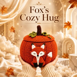May include: A round, orange, crocheted pumpkin-shaped container with a lid, featuring a dark brown stem and green leaves. A small, crocheted fox with white, orange, and black details is attached to the front. The text Fox's Cozy Hug is above.