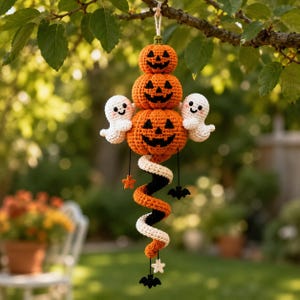 May include: A hanging crochet decoration featuring three stacked orange pumpkins with black stitched faces, flanked by two small white ghosts. Below the pumpkins is a spiraling tail in orange, black, and white, accented with small stars and bats.