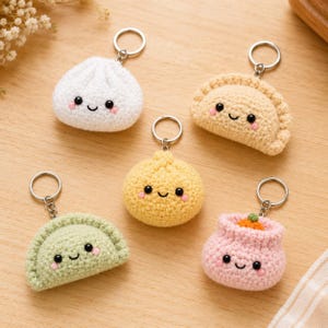 May include: Five crochet keychains shaped like various dumplings and buns. Each item features a soft yarn texture, black bead eyes, a stitched smile, and pink blush marks. The set includes white, beige, yellow, light green, and pink colors.