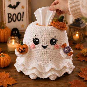 May include: A white crocheted ghost tissue box cover featuring a ruffled base, black button eyes with eyelashes, pink cheeks, and a small smile. The ghost holds a small orange crocheted pumpkin in one hand and a purple candy in the other.