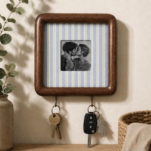May include: A square dark wood wall-mounted key holder featuring a central black and white photograph of two people kissing. The frame is backed by a light blue and white striped fabric and includes two metal hooks holding two sets of keys.