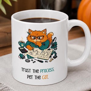 May include: A white ceramic mug filled with dark coffee, featuring a graphic of an orange cat wearing glasses and a teal shirt, working with blueprints, tools, and a calculator. Below the illustration, the text reads: Trust the process. Pet the cat.
