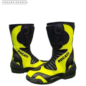 Peut inclure: Une paire de bottes de moto jaune fluo et noir avec le mot DUCATI écrit en noir sur le côté. Les bottes présentent des panneaux de protection noirs au niveau du tibia, de la cheville et des orteils, avec des zones flexibles en accordéon.
