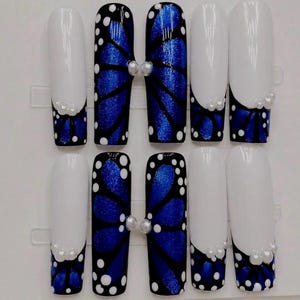 Op de afbeelding: Een set van tien press-on nagels met een lang, vierkant ontwerp. De nagels tonen een combinatie van effen witte uiteinden en een levendig blauw vlindervleugelpatroon met zwarte aders en witte stippen, geaccentueerd met kleine witte parels.