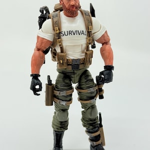Könnte beinhalten: Eine bärtige Actionfigur mit rotbraunem Haar, die ein grünes Stirnband, ein weißes T-Shirt mit dem in Schwarz gedruckten Wort SURVIVAL, eine beige taktische Weste mit Taschen, eine olivgrüne Tarnhose, schwarze Handschuhe und schwarze Stiefel trägt.