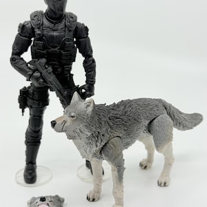 May include: A black tactical action figure wearing a mask and holding a small firearm, standing next to a grey and white wolf figurine. A separate wolf head piece with an open mouth showing teeth is positioned on the ground in front of the figures.