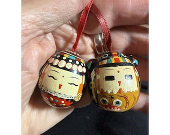 May include: Two small, round, wooden ornaments featuring hand-painted faces and decorative patterns. One ornament displays a figure with closed eyes and a red headpiece, while the other shows a figure with a tiger-like design. Both have red ribbons.