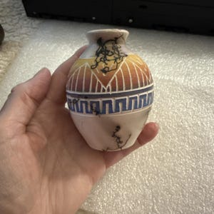 Puede incluir: Un pequeño jarrón de cerámica de color blanco roto con forma redondeada, que presenta una banda decorativa con patrones de rayos de sol en naranja y amarillo sobre un diseño geométrico azul de grecas. La superficie tiene finas líneas negras.