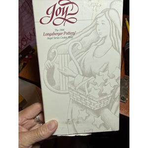 Puede incluir: Una caja rectangular blanca con un logotipo granate que dice Joy. Debajo, el texto dice The 1996 Longaberger Pottery Angel Series Cookie Mold. La caja muestra un dibujo lineal de un ángel sosteniendo un arpa y una cesta llena de estrellas.