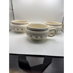 May include: Three cream-colored ceramic soup bowls with handles, each featuring a thin maroon horizontal band near the rim and a repeating pattern of small, dark maroon heart-shaped floral motifs around the lower body of the bowls.