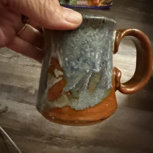 Puede incluir: Una taza de cerámica con un asa gruesa y curva, que presenta un esmalte texturizado que transita desde un naranja terracota profundo en la base hasta un acabado moteado en tonos azul grisáceo hacia el borde, con un aspecto rústico.