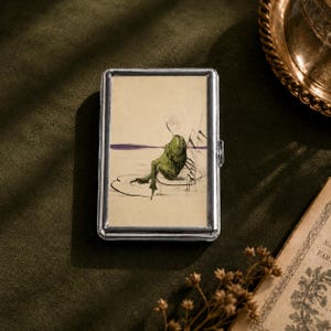 May include: A rectangular silver-toned metal case featuring a printed illustration of a green frog sitting on a lily pad. The artwork depicts the frog in a relaxed posture against a light beige background with a thin purple horizontal line.