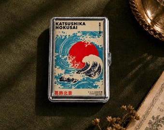 Könnte beinhalten: Ein rechteckiges Etui aus silberfarbenem Metall mit einem grafischen Druck nach einem Kunstwerk von Katsushika Hokusai. Das Design zeigt eine große rote Sonne hinter stilisierten blauen und weißen Meereswellen. Der Text lautet Katsushika Hokusai.