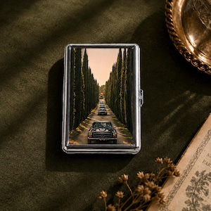 May include: A rectangular silver metal cigarette case featuring a printed image of a black Mercedes-Benz car driving down a long driveway lined with tall, slender cypress trees under a warm, golden-toned sky at sunset.
