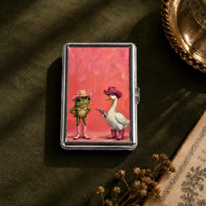 May include: A rectangular silver metal case featuring a printed illustration of a green frog and a white goose standing against a pink background. Both figures wear pink cowboy hats and boots, and the goose holds a small pink toy revolver.