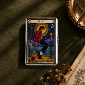 May include: A rectangular silver metal cigarette case featuring a colorful tarot card illustration. The design depicts a figure in a red jacket and black pants, with the text BEAT IT at the top, set against a starry night sky, mountains, and water.