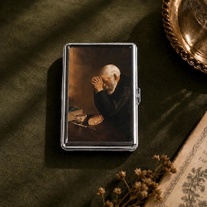 May include: A rectangular silver-toned metal case featuring a printed reproduction of a painting depicting an elderly person with a white beard praying at a table with a loaf of bread, a small bowl, and a book in a dark, warm-toned setting.