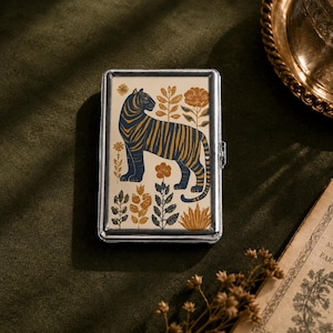 May include: A rectangular metal case with a silver-toned frame and a cream-colored top panel. The panel features a dark blue tiger with orange stripes standing among stylized orange and blue floral and botanical illustrations.