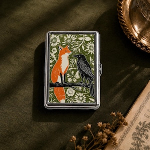 Pode incluir: Um estojo retangular de metal prateado com um design impresso de uma raposa laranja e um corvo preto pousados num ramo sobre um fundo verde com padrões florais brancos. O estojo possui uma moldura metálica e um mecanismo de dobradiça.