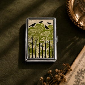 May include: A rectangular silver metal case featuring a graphic print of black birds perched in a forest of green and white trees. The case has a polished metallic frame and a hinged closure, displaying a detailed illustration of woodland scenery.