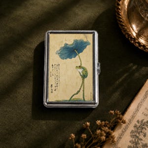 May include: A rectangular silver metal case featuring a printed illustration of a green frog perched on a stem beneath a large blue lotus leaf. The background is a textured cream color with black Japanese calligraphy characters on the left side.