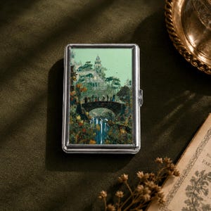 May include: A rectangular silver metal case featuring a printed illustration of a group of figures crossing a stone bridge over a waterfall, with a tall building in the background. The case has a polished metallic frame and a hinged side closure.