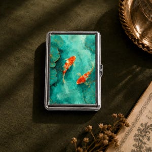 Peut inclure: Un étui à cigarettes rectangulaire en métal orné d'une illustration vibrante de deux poissons koï orange et blancs nageant dans une eau bleu sarcelle profonde parmi des nénuphars. L'étui possède un cadre argenté et une fermeture à charnière.