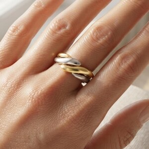 May include: A multi-tone ring featuring three intertwined bands in polished yellow gold, rose gold, and silver tones. The ring has a smooth, rounded, and fluid design that wraps around the finger, creating a layered and cohesive appearance.