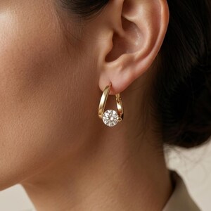 May include: A gold hoop earring featuring a teardrop-shaped frame with a single round, clear, faceted gemstone suspended at the base. The metal has a polished finish, and the earring is secured through the earlobe with a hinged closure mechanism.