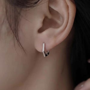 May include: A small, silver-toned hoop earring with a distinct hexagonal shape. The earring features a polished, metallic finish and a hinged closure mechanism, designed to sit snugly against the earlobe with a clean, geometric aesthetic.
