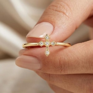 Peut inclure: Une bague en or dotée d'un petit motif central en forme de croix orné de cinq pierres claires et scintillantes. L'anneau est fin et poli, présentant un design délicat qui se place horizontalement sur le doigt avec une finition métallique raffinée.