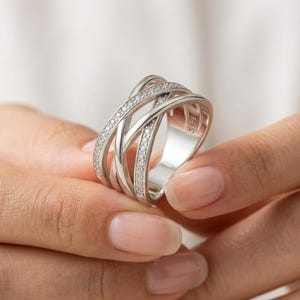 Puede incluir: Un anillo de tono plateado con un diseño cruzado de múltiples capas formado por bandas delgadas. Las bandas exteriores están incrustadas con pequeñas piedras claras y brillantes. El interior del anillo tiene grabado el texto S925, indicando su composición.