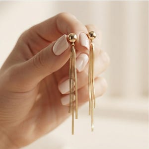 May include: A pair of gold-tone earrings featuring a small polished sphere stud at the top, from which three delicate, flat, flexible chain tassels hang down in a vertical arrangement, creating a shimmering and fluid metallic appearance.