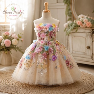 May include: A strapless dress in a light beige hue featuring a full, layered tulle skirt. The bodice and skirt are adorned with multi-colored fabric flowers in shades of pink, blue, yellow, and purple, accented with sequins and embroidery.