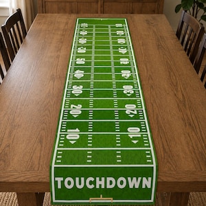May include: A rectangular green table runner featuring a white graphic print of an American football field with yard lines, numbers, and the word TOUCHDOWN printed in bold white capital letters at the bottom end of the field design.