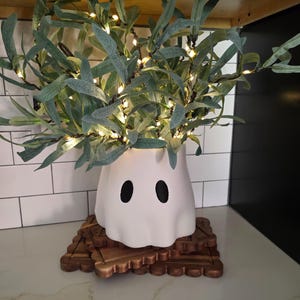 May include: A white ceramic ghost-shaped planter featuring two black oval eyes, filled with green leafy branches intertwined with small warm white string lights, resting on a dark brown wooden scalloped-edge square base.