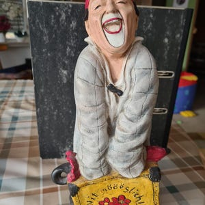 Könnte beinhalten: Eine Keramikfigur einer lachenden Person mit roter Mütze und hellgrauem, strukturiertem Mantel. Die Figur steht hinter einem gelben rechteckigen Schild mit dem schwarzen Text Nit mööööglich! und drei kleinen roten Blumenillustrationen.