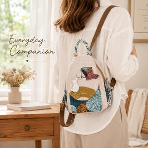 May include: A small beige canvas backpack featuring a braided handle and an embroidered design of a white cat sitting on a textured arch. The lower section displays a blue and orange abstract pattern, complemented by brown straps and metal zippers.