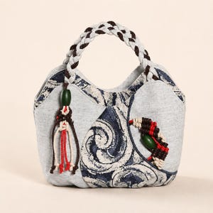 Può includere: Una borsa con corpo in tessuto grigio chiaro e motivi a vortice blu scuro e bianchi. La borsa presenta un manico intrecciato bianco e marrone scuro, una nappa decorativa con una perla verde e un inserto a mezzaluna in corda rossa e marrone.