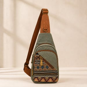 May include: A sage green canvas sling bag featuring a brown leather-like strap and trim. The front pocket displays a triangular panel with a geometric pattern in black, white, blue, and brown. It includes metal zippers and a D-ring attachment.