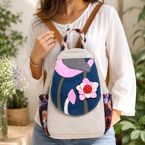 May include: A beige backpack featuring a dark blue flap with pink and white floral applique, a braided handle, and side pockets with a dark floral pattern. The bag has brown shoulder straps and a quilted gray accent on the top of the flap.