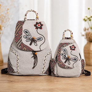 May include: Two light gray backpacks of different sizes featuring a braided top handle, a flap closure with a dragonfly and flower embroidery, and side panels with a geometric pattern in red, black, and white. Each bag has a drawstring closure.