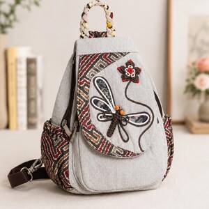 May include: A light gray backpack featuring a braided top handle and a front flap decorated with an embroidered dragonfly and flower motif. The bag includes side panels with a geometric pattern in red, brown, and white, along with dark brown straps.