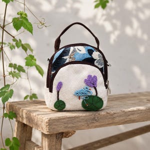 May include: A small, rounded handbag featuring a cream-colored canvas front with embroidered purple flowers and a blue bird on green lily pads. The upper section has a blue and white floral pattern, finished with dark brown trim and a top handle.